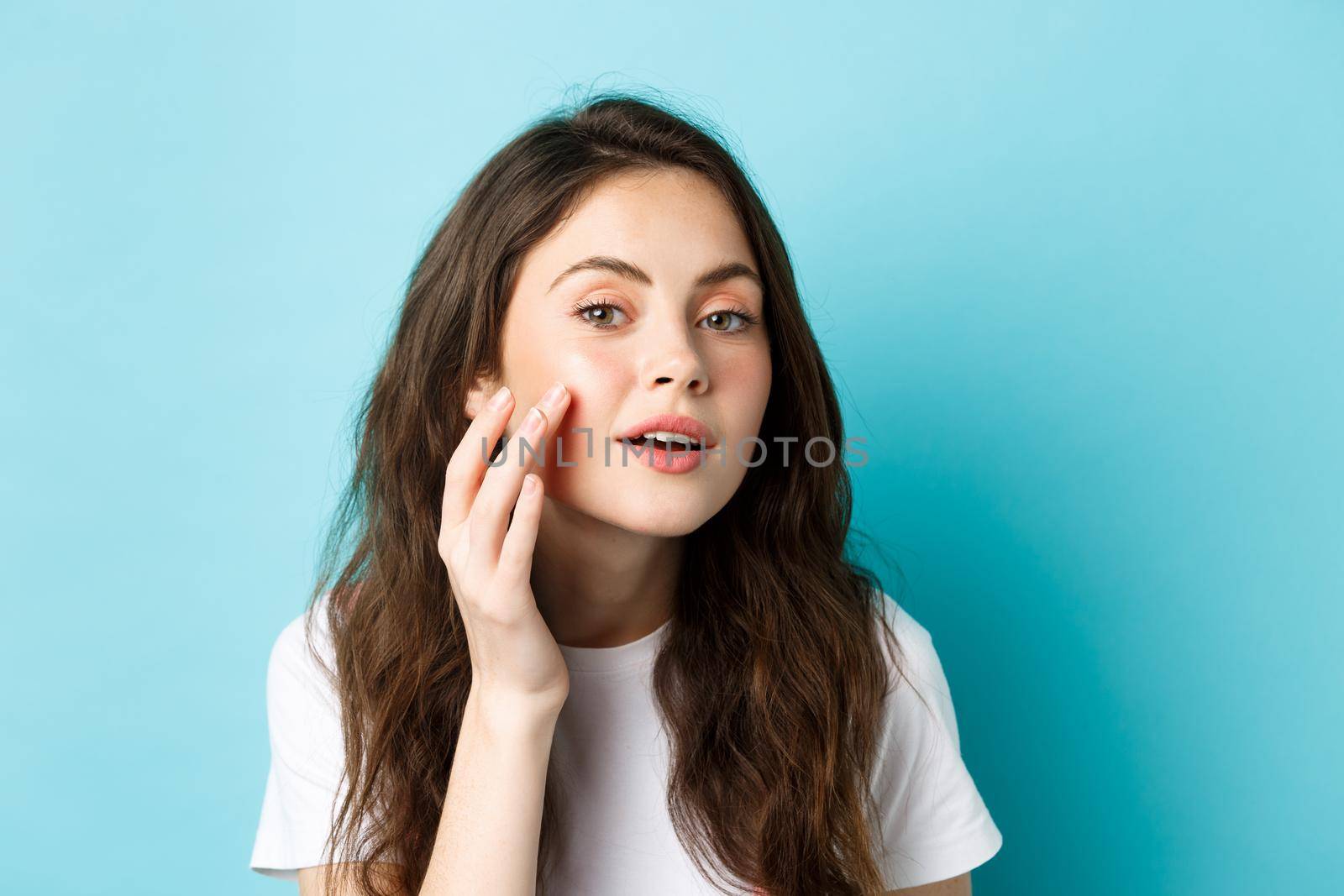 Skin care. Close up of young woman looking in mirror and touching clean glowing facial skin, checking out cosmetics after effect, apply beauty product, standing over blue background.