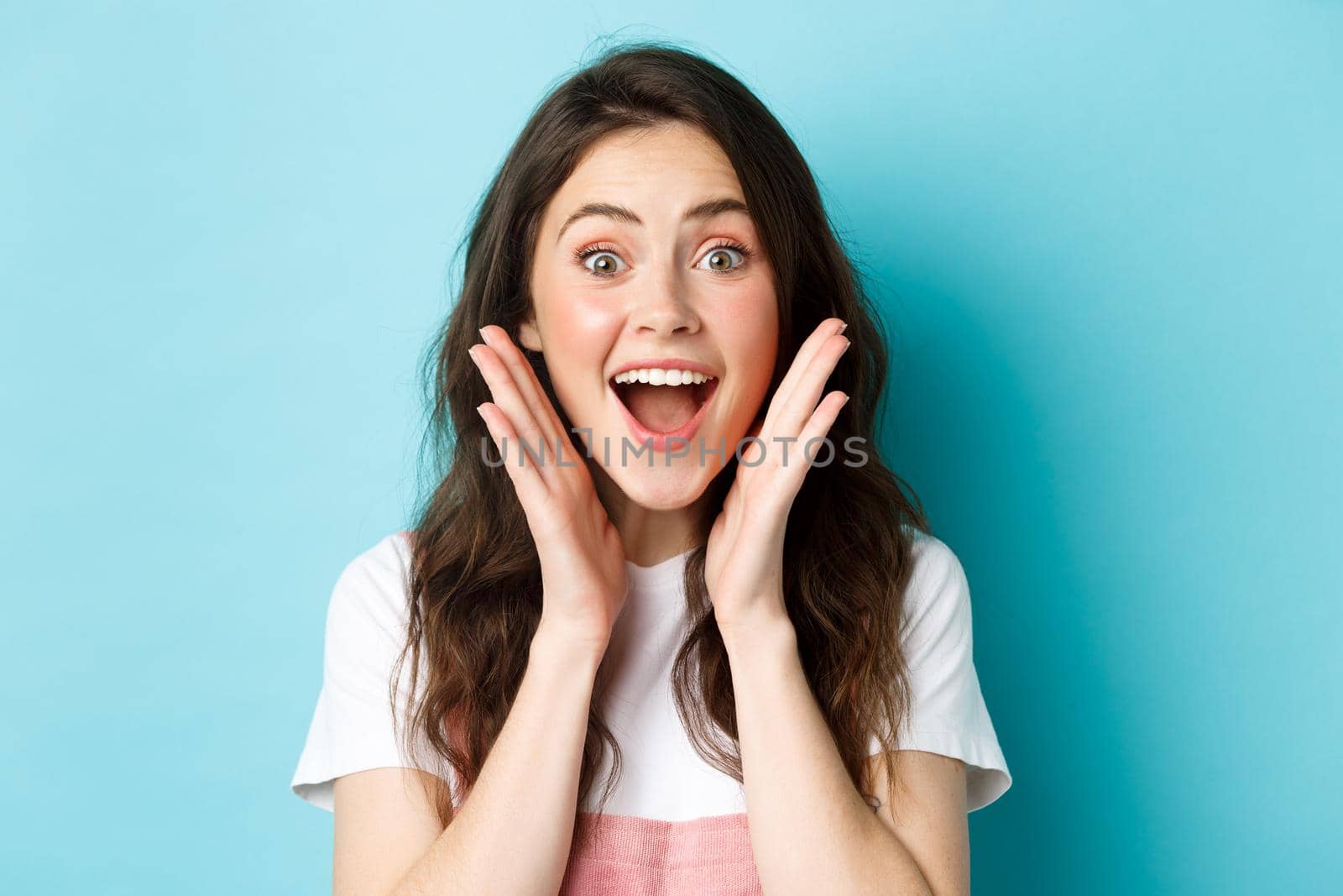 Close up of excited young woman holding hands near face, gasping and looking amazed at camera, stare surprised at super cool news, standing over blue background.