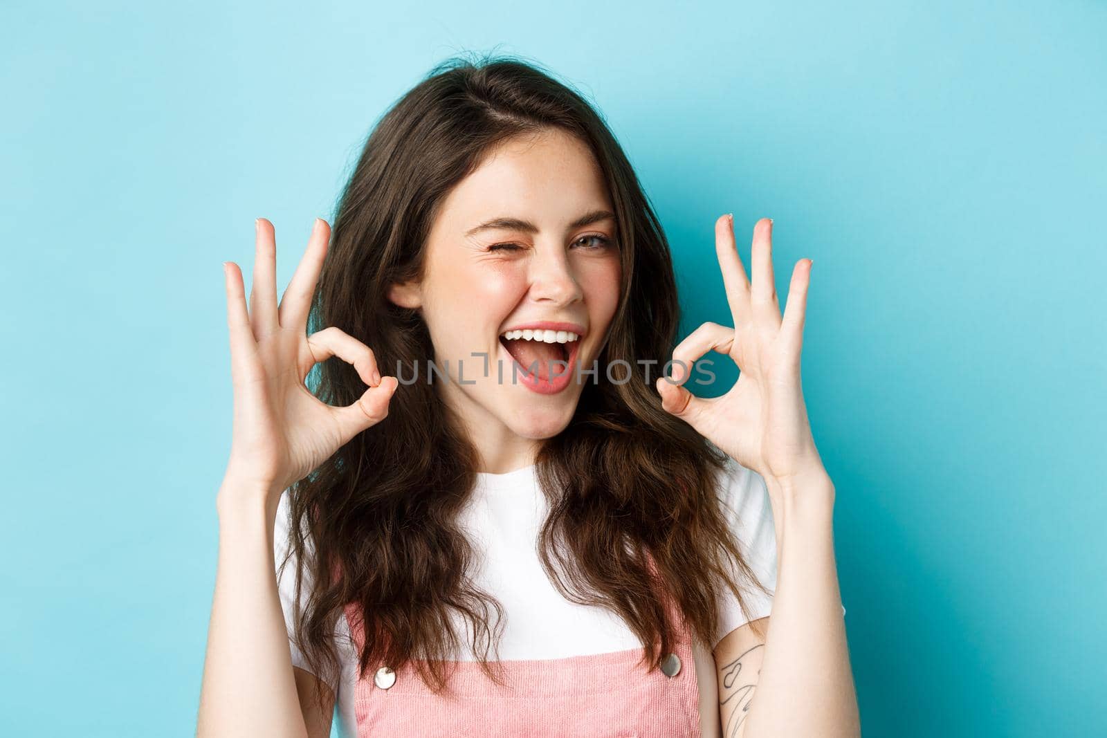 Close up of cheerful caucasian woman agree and say yes, make alright okay sign and winking assuringly at camera, standing satisfied over blue background.