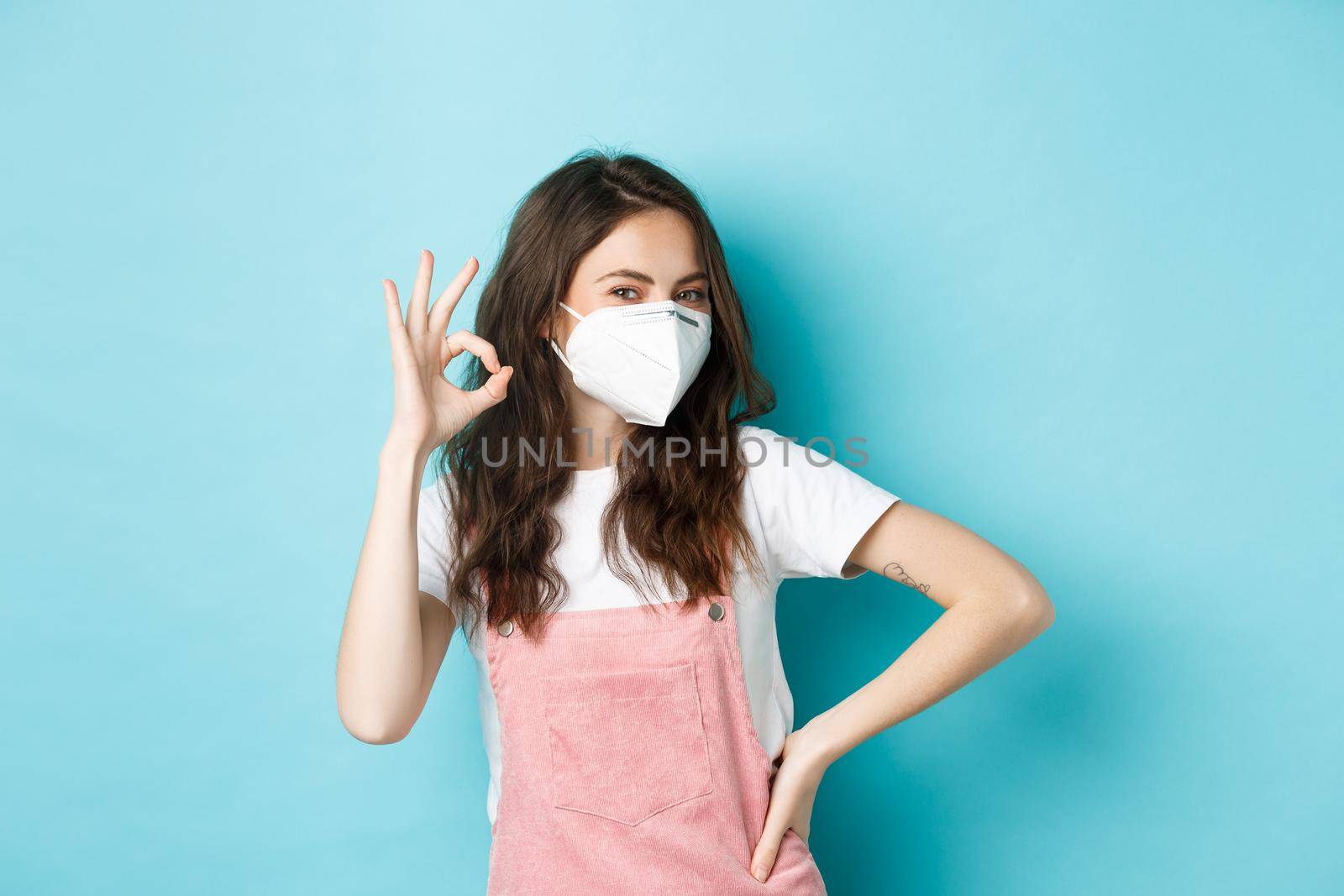 Covid, health and pandemic concept. Very good. Young supportive woman wearing medical respirator and showing okay sign in approval, praise wearing face masks in public, blue background.