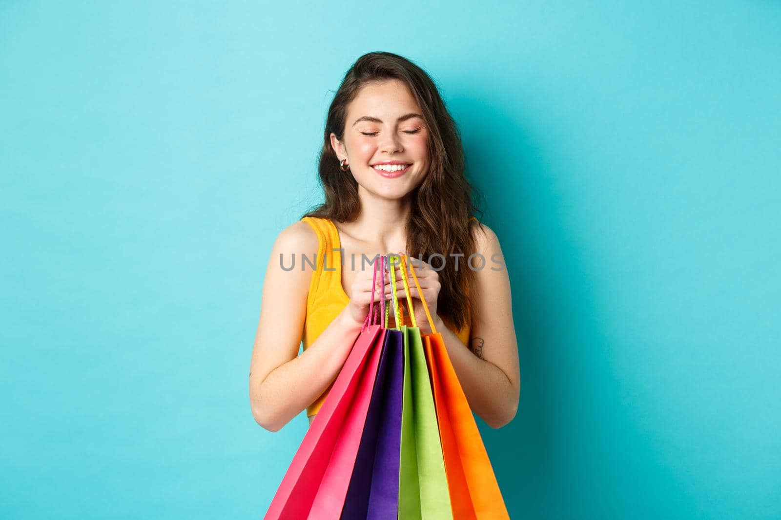 Young beautiful woman daydreaming about wearing new clothes, holding shopping bags, close eyes and smile dreamy, standing over blue background.