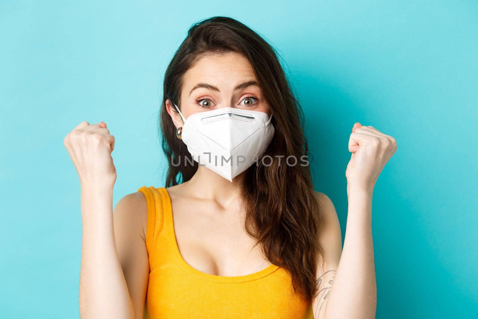 Health, covid-19 and lockdown concept. Excited young woman in respirator mask, saying yes, express rejoice and triumph, making fist pump from good news, standing against blue background.