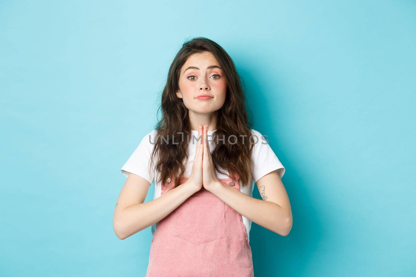 Please help me. Cute brunette woman asking for favour, holding hands in pray, begging you to buy something, standing against blue background.