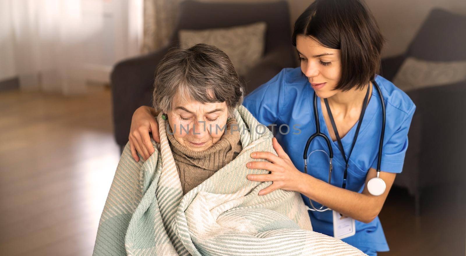 A young nurse takes care of an elderly 80-year-old woman at home, wraps a blanket around her. Happy retired woman and trust between doctor and patient. Medicine and healthcare.