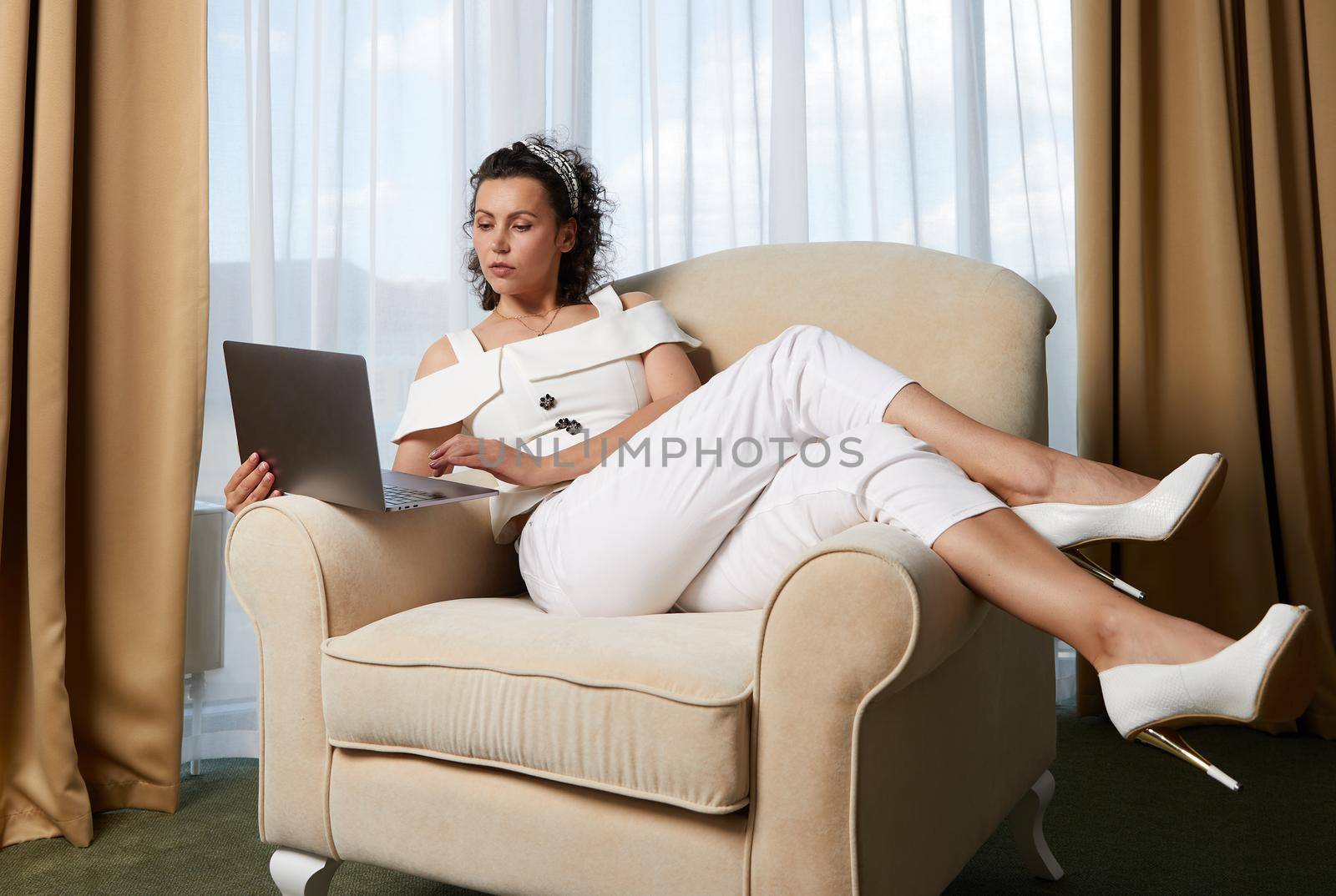 Beautiful young woman using laptop sitting in armchair. Concept of relaxed business lady