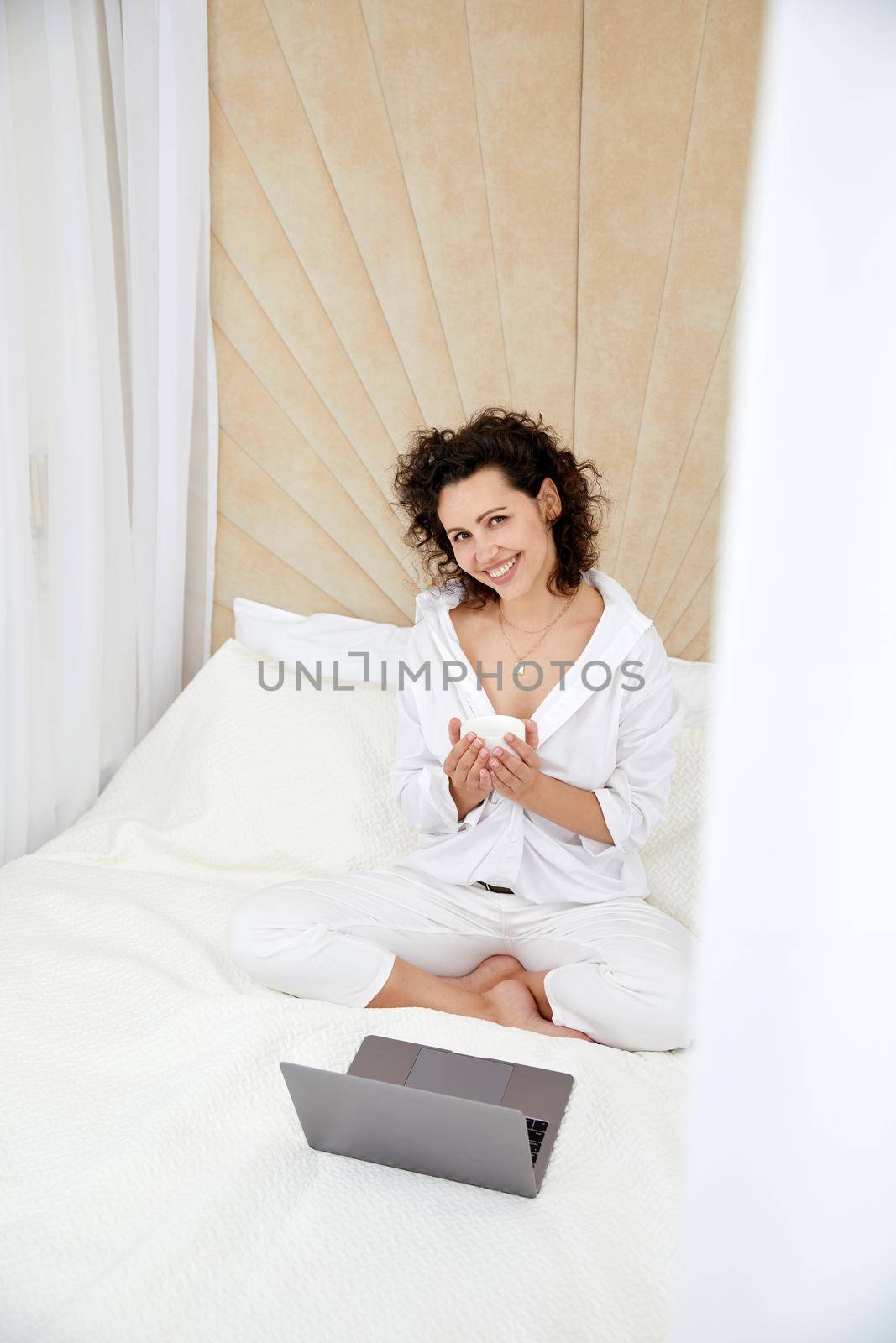 Curly girl using laptop computer sitting on white bed browsing or chatting with friends online by Mariakray