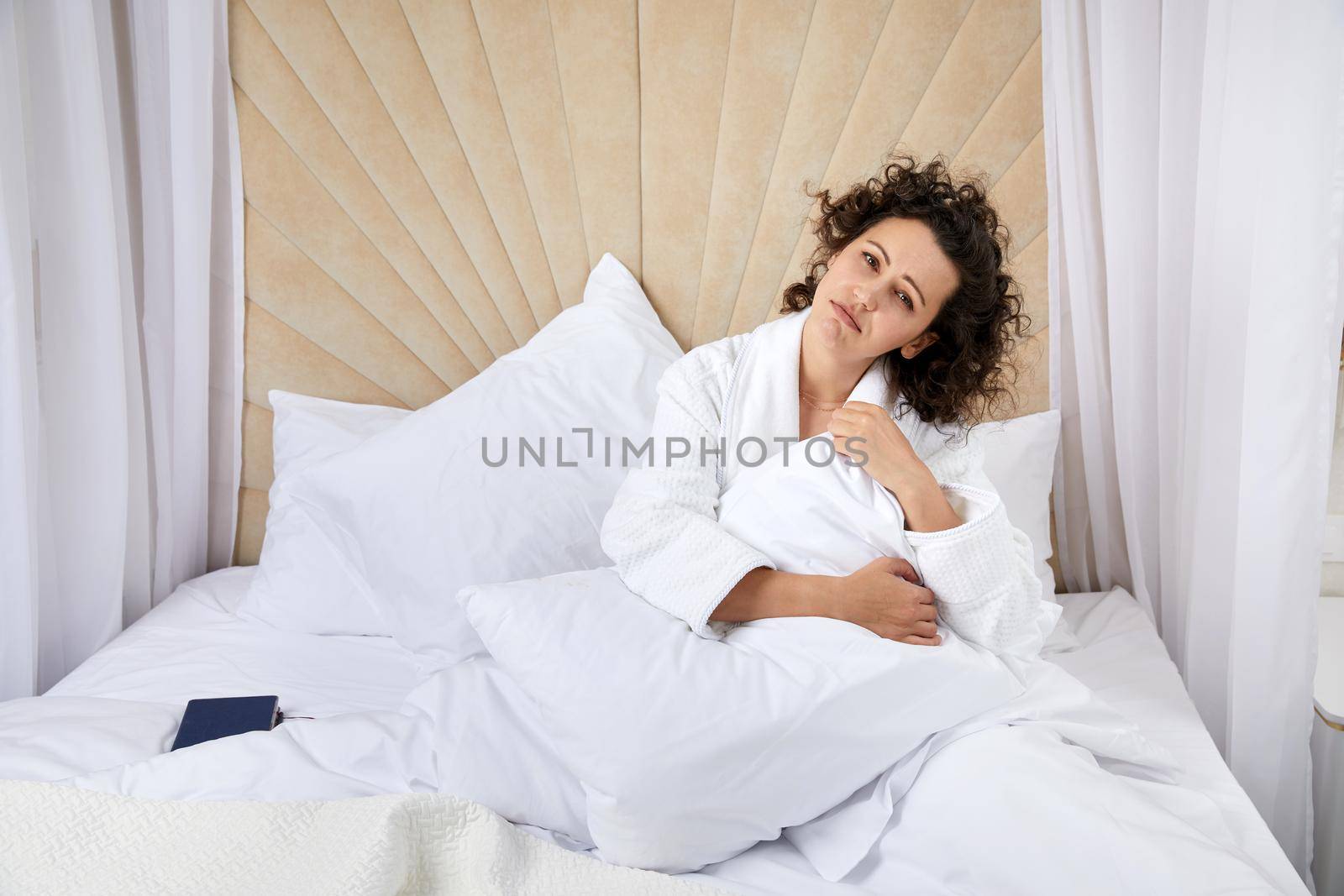 Sad depressed curly woman hugging pillow sitting in white bed alone