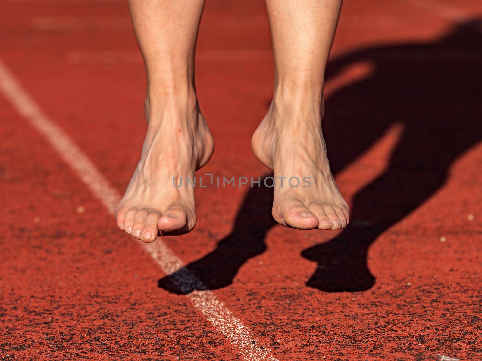 Middle aged barefoot jumping woman warm up muscles and stretching body on stadium run track. Real people do sport