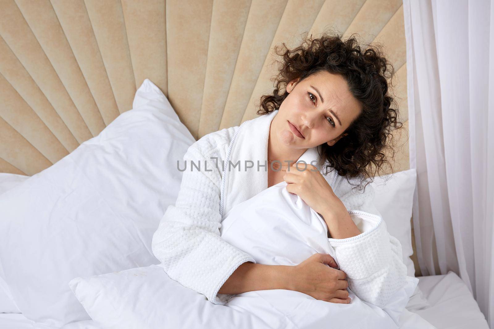 Sad depressed curly woman hugging pillow sitting in white bed alone