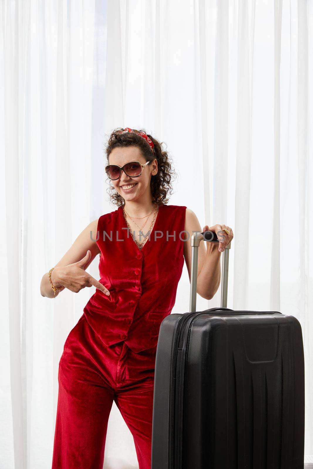 Glamour young woman wearing red velvet suit standing smiling and pointing on suitcase in hotel room. Travel concept by Mariakray