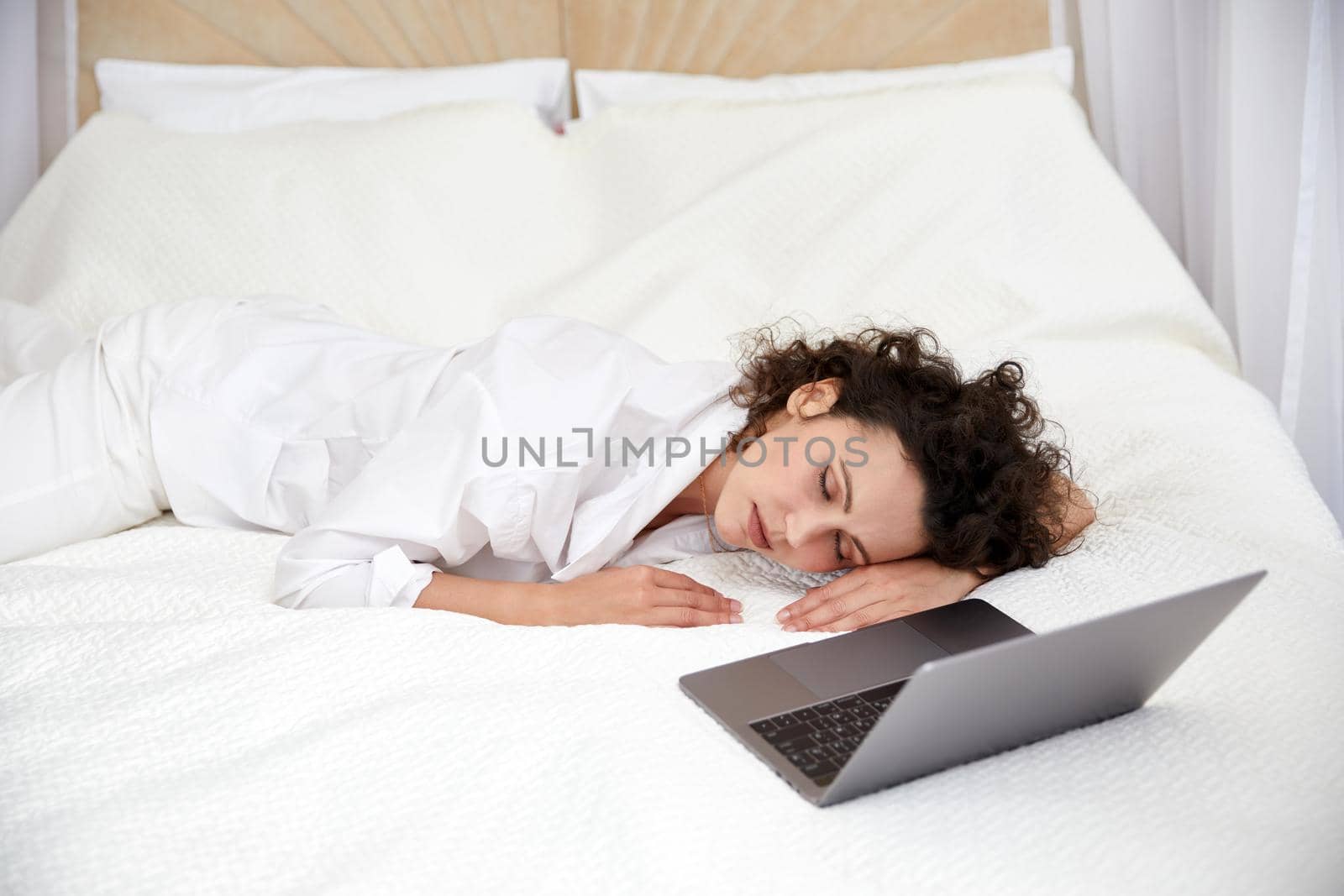 Curly girl sleeping tired after using laptop computer on white bed browsing and chatting with friends online