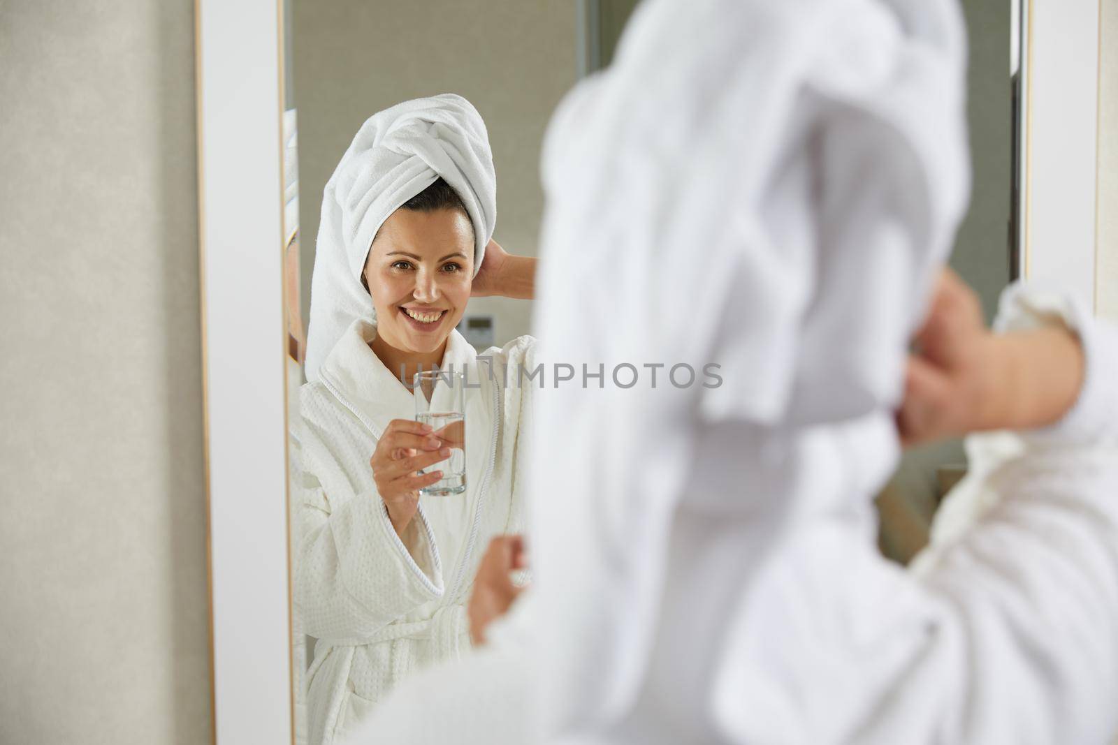 Beautiful happy girl drinking and holding glass of water standing in front of mirror in the morning. Concept of healthy lifestyle