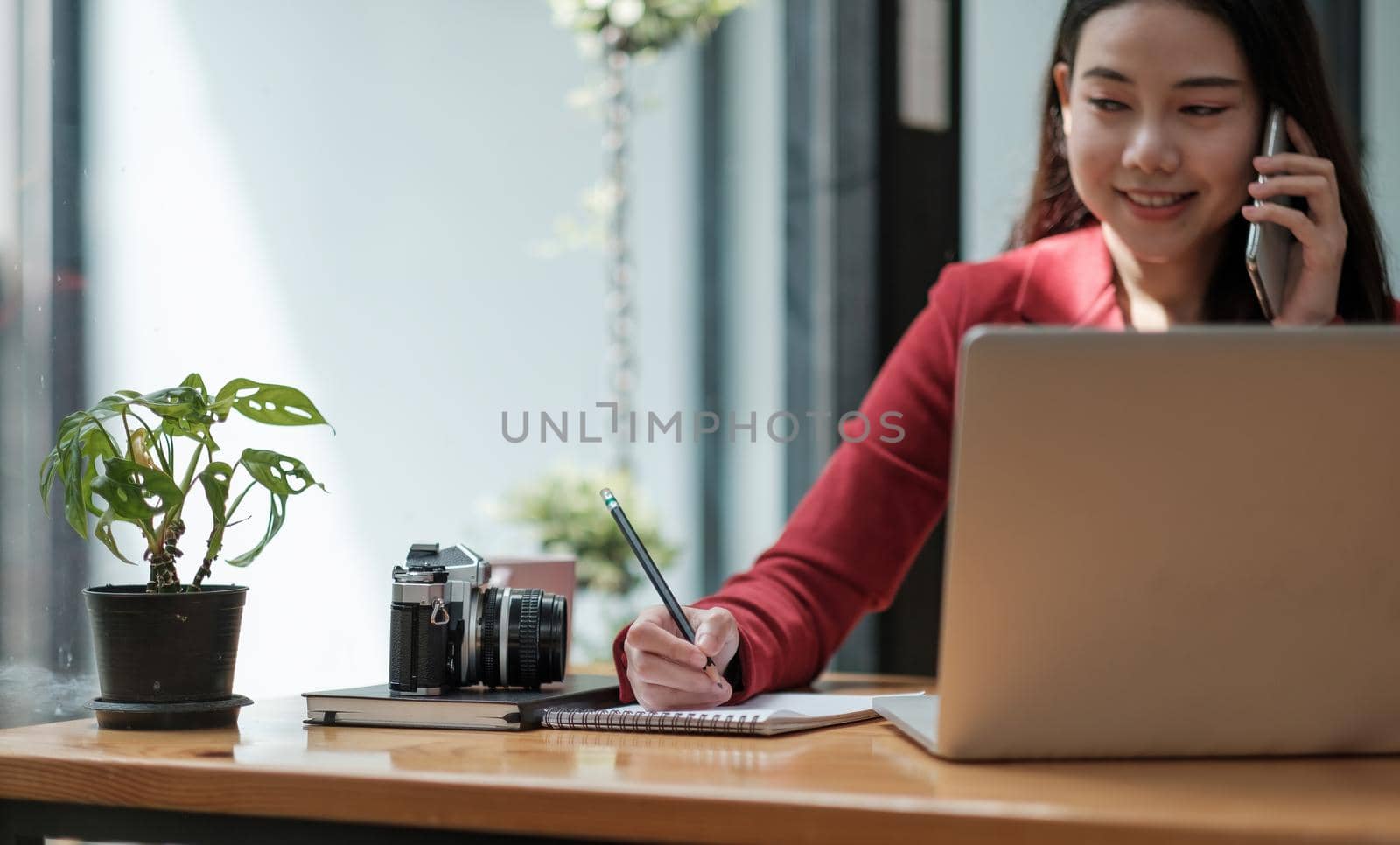 Young woman talking on mobile phone and writing notes while sitting at her desk. Asianfemale working in home office.