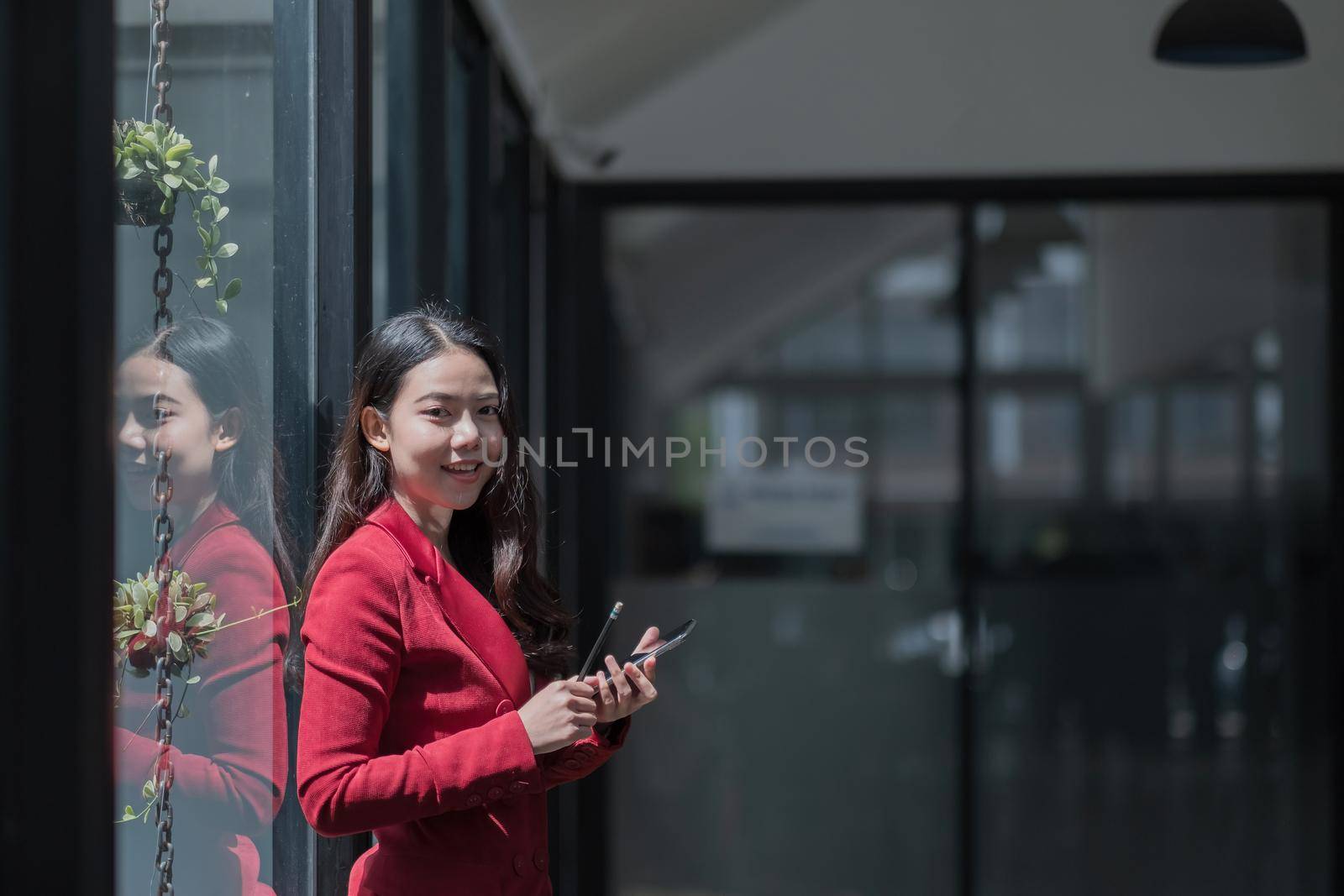 Image of young asian woman holding mobile phone while working in office, she looking at camera by nateemee