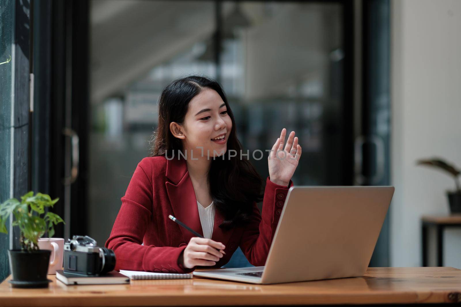 Image of happy asian woman taking note and waving hand at laptop, while speaking or chatting on video call in office.