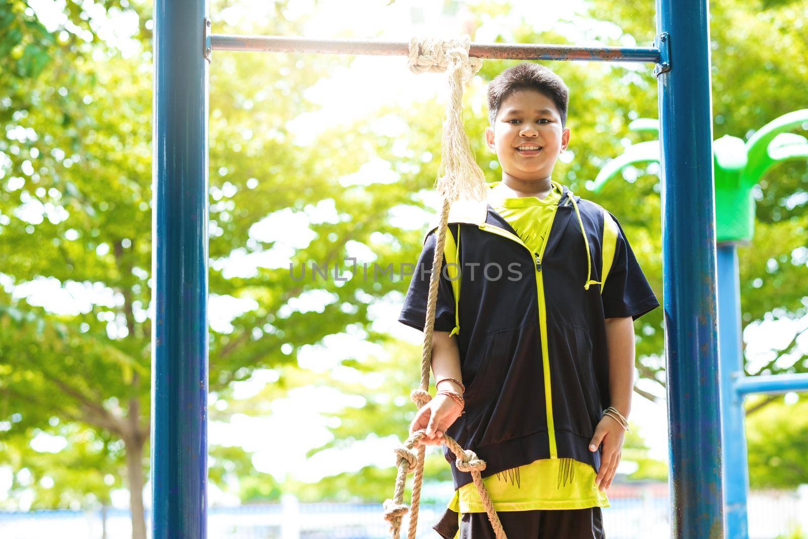 Young asian boy hang the yellow bar by his hand to exercise at out door playground under the big tree. by Benzoix