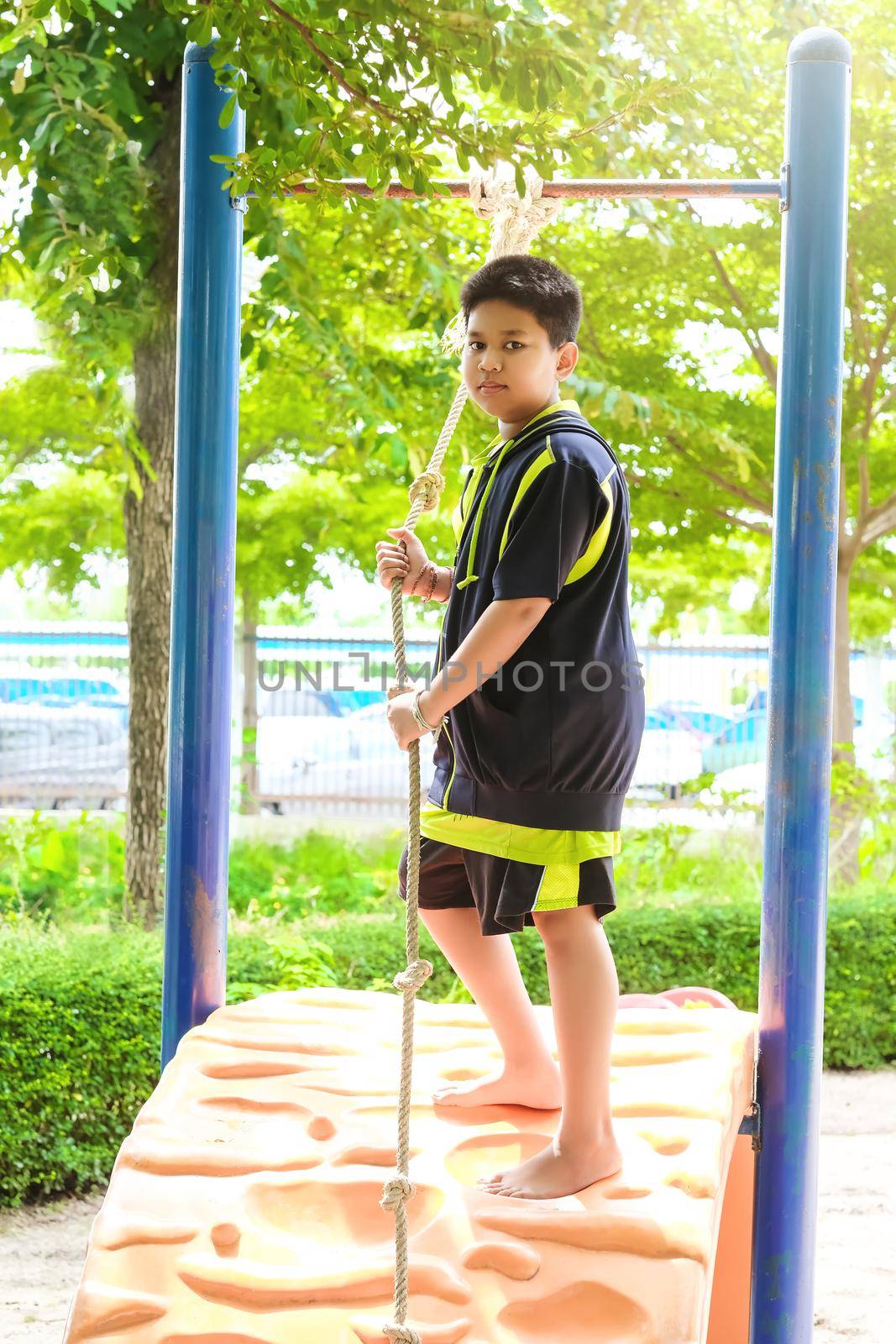 Young asian boy hang the yellow bar by his hand to exercise at out door playground under the big tree. by Benzoix