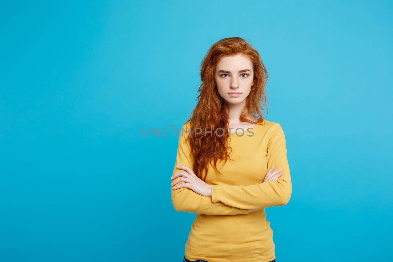 Portrait of young beautiful ginger woman with tender serious face crossing arms looking at camera. Isolated on pastel blue background. Copy space. by Benzoix