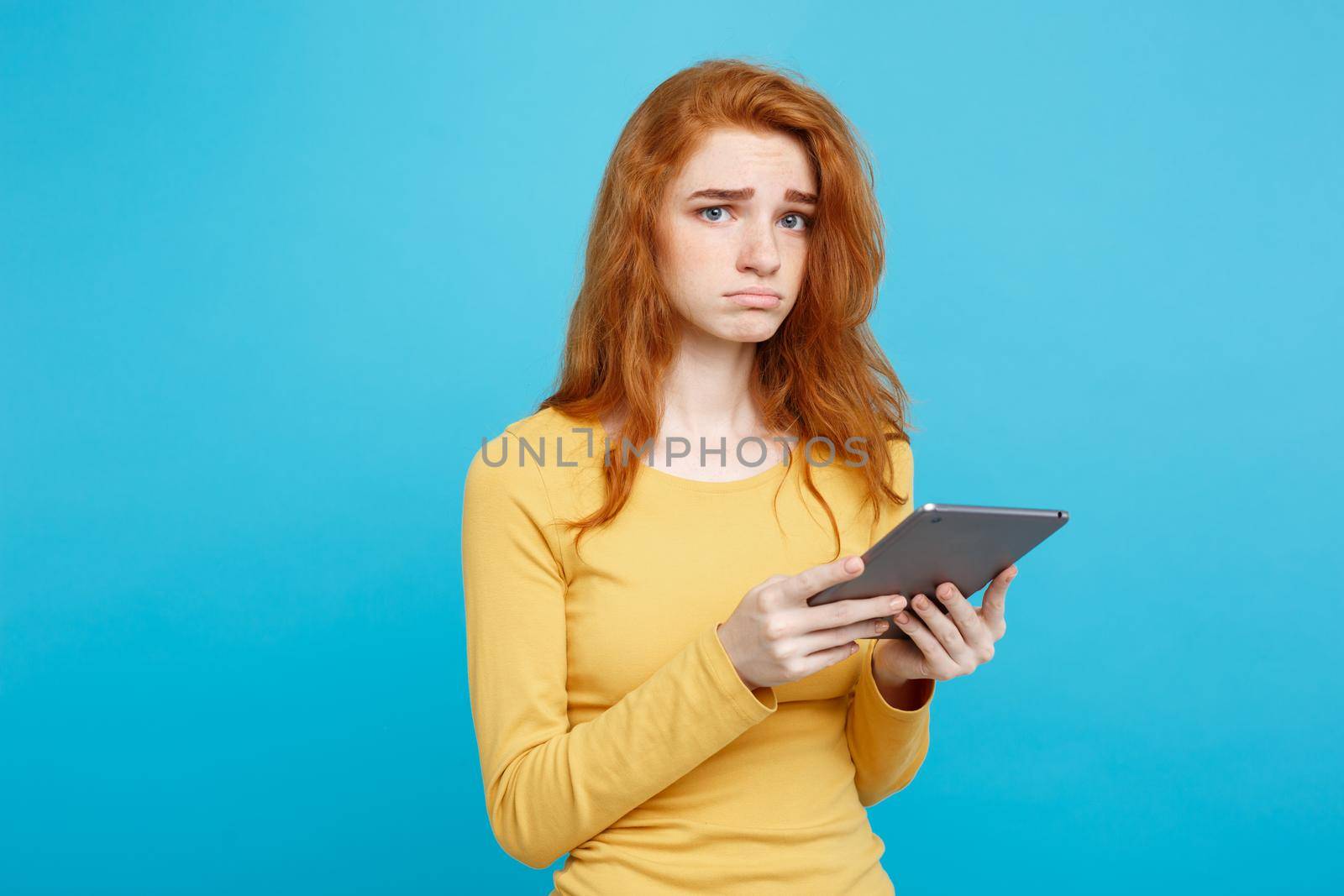 People and Technology Concept - Close up Portrait young beautiful attractive tender ginger redhair girl shocking and sad on digital table with wining something. Blue Pastel Background. Copy space. by Benzoix
