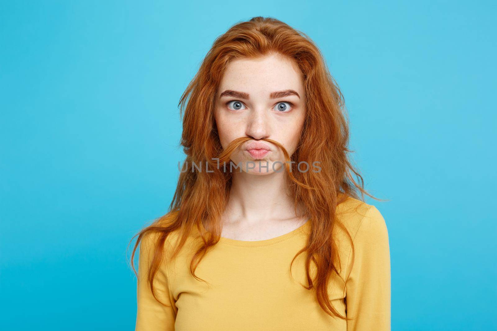 Headshot Portrait of happy ginger red hair girl imitating to be man with hair fake mustache.Pastel blue background. Copy Space.