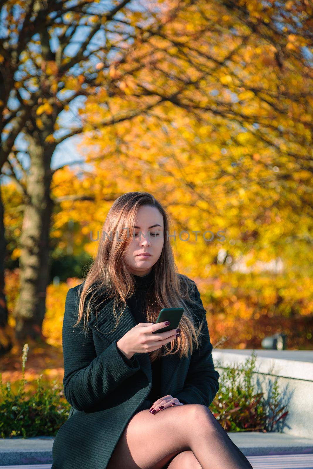 A woman with long hair sits on a bench in an autumn park and looks into a mobile phone. by Yurich32