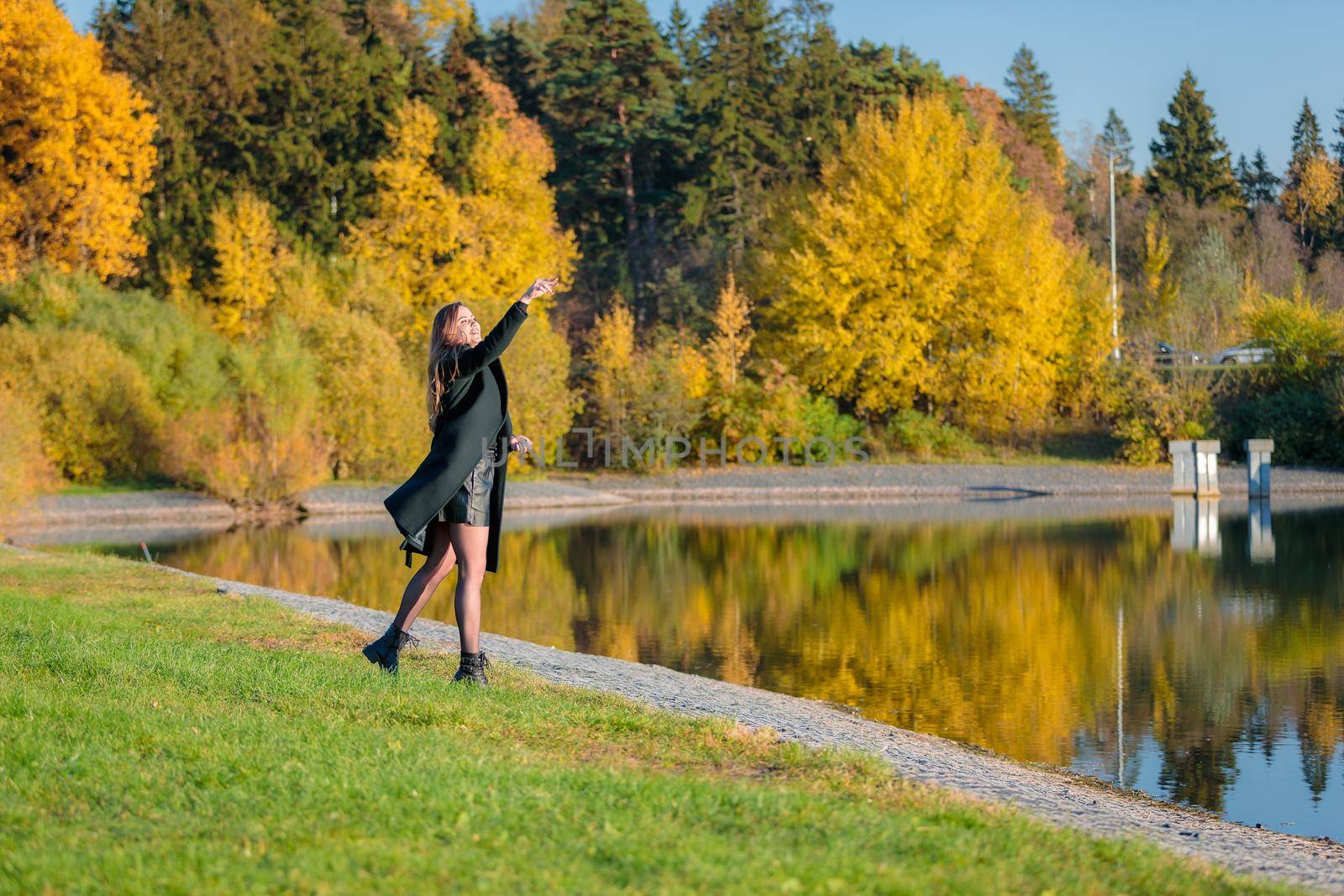 A woman with long hair walks in an autumn park by a pond and throws stones into the water. Autumn season.