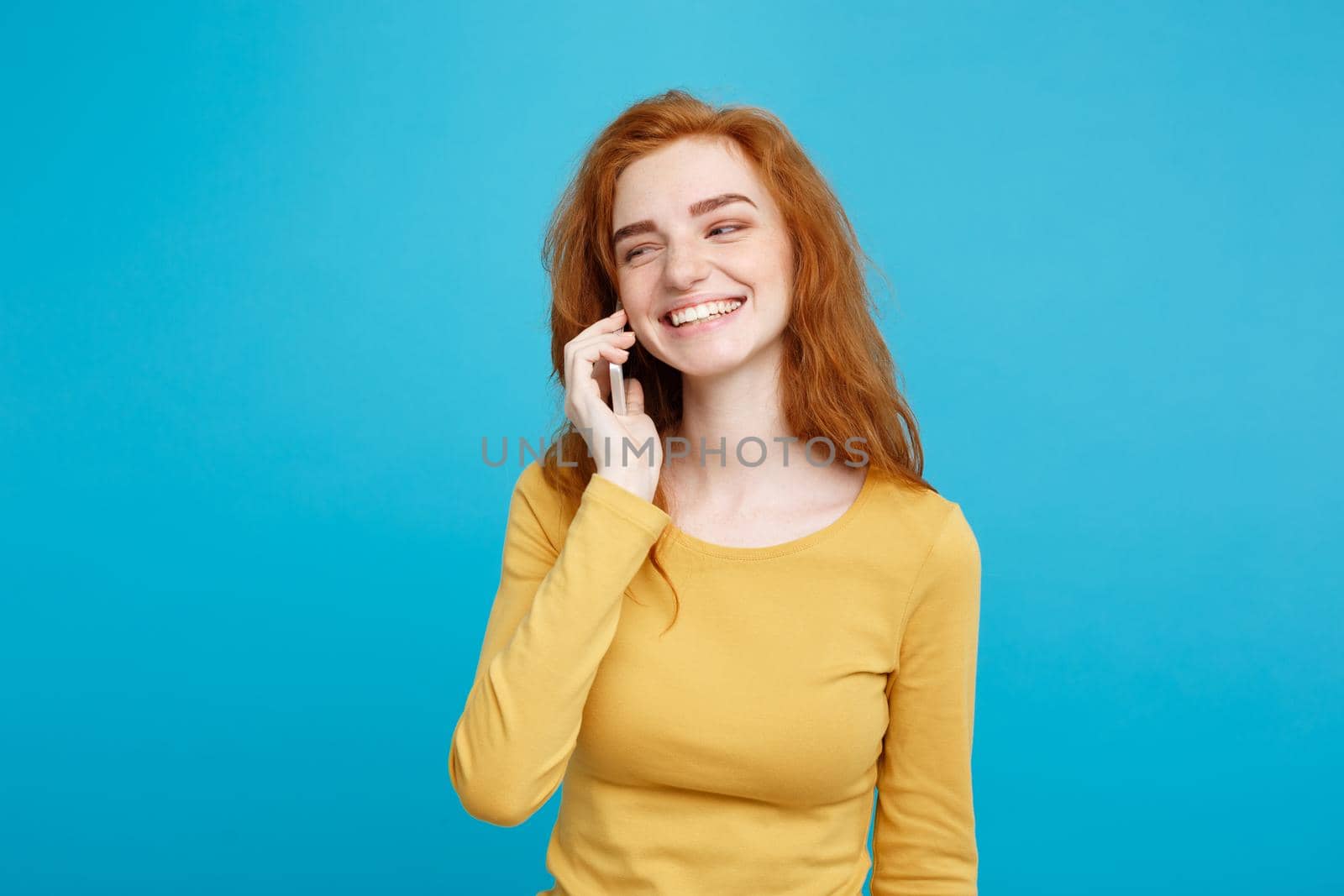 Lifestyle and Technology concept - Portrait of cheerful happy ginger red hair girl with joyful and exciting talking with friend by mobile phone. Isolated on Blue Pastel Background. Copy space.