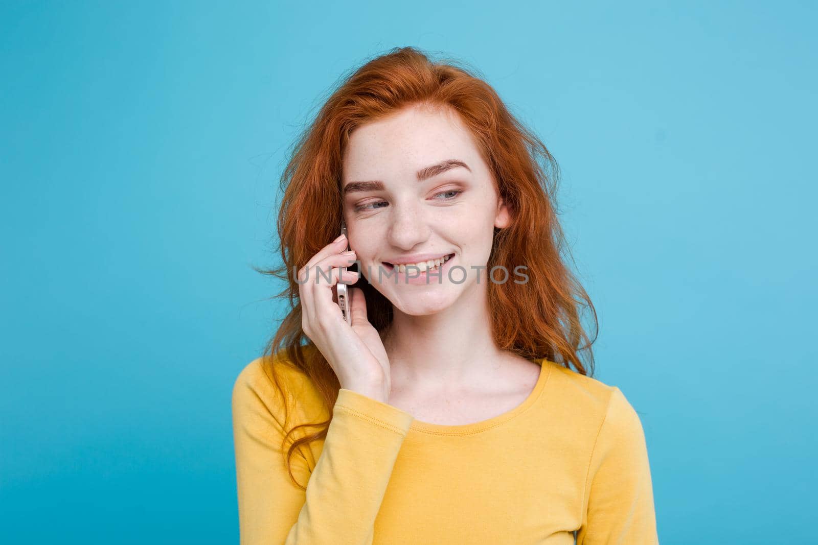 Lifestyle and Technology concept - Portrait of cheerful happy ginger red hair girl with joyful and exciting talking with friend by mobile phone. Isolated on Blue Pastel Background. Copy space.