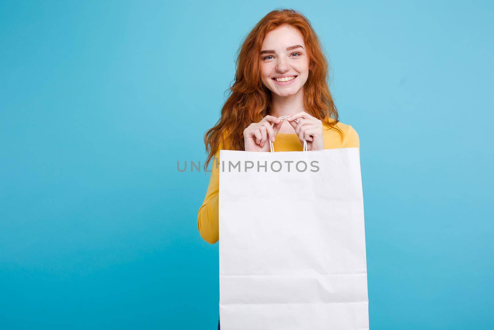 Shopping Concept - Close up Portrait young beautiful attractive redhair girl smiling looking at camera with white shopping bag. Blue Pastel Background. Copy space by Benzoix