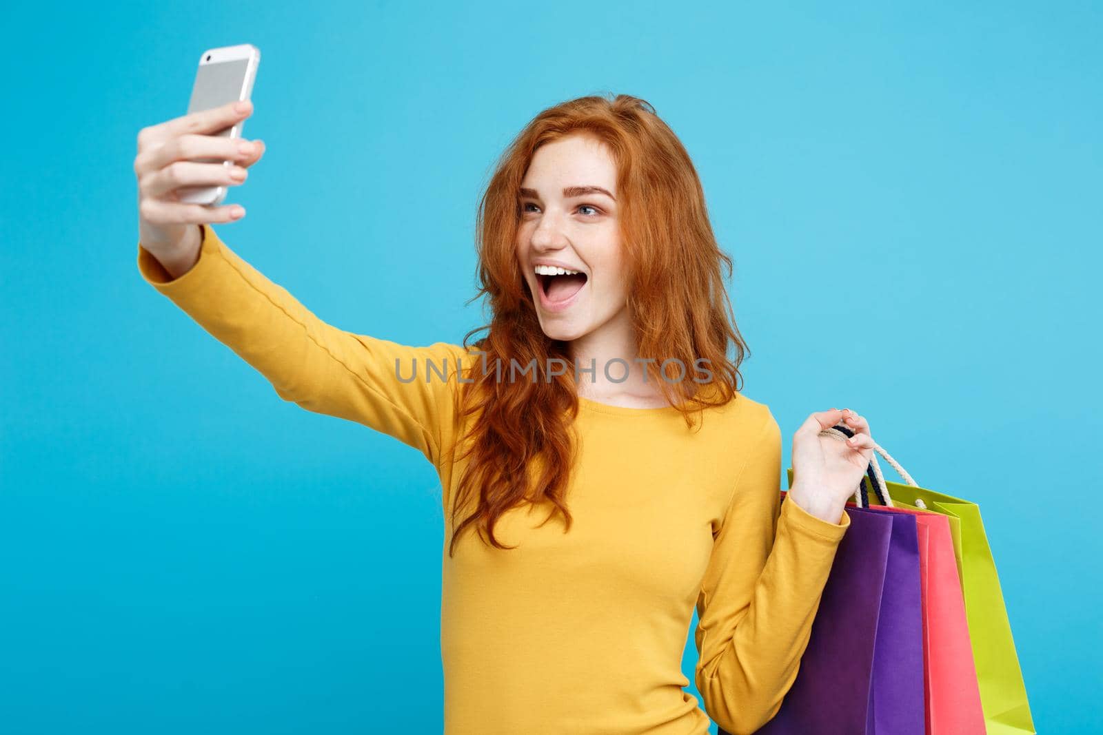 Shopping Concept - Close up Portrait young beautiful attractive redhair girl smiling looking at camera with white shopping bag and selfie. Blue Pastel Background. Copy space.