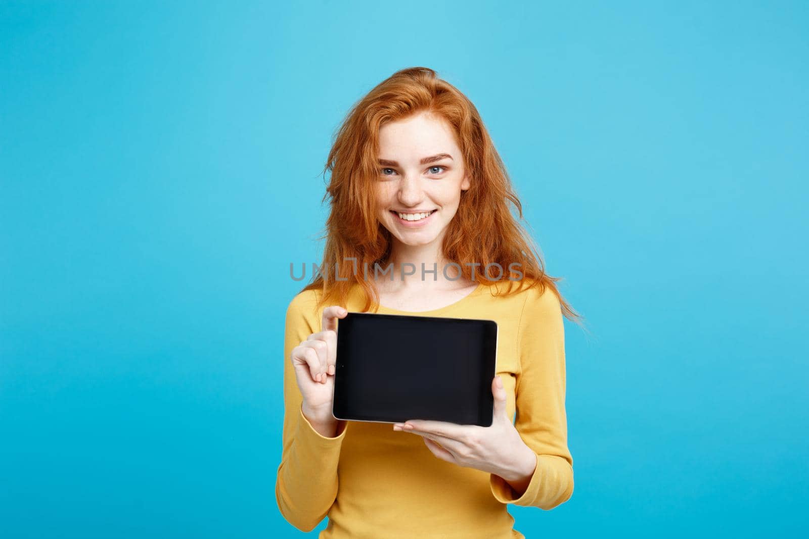 Close up Portrait young beautiful attractive redhair girl smiling showing digital tablet screen on black. Blue Pastel Background.