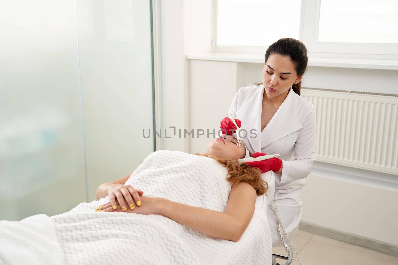 Beautiful Woman At Spa Clinic Receiving Stimulating Electric Facial Treatment From Therapist. Closeup Of Young Female Face During Microcurrent Therapy
