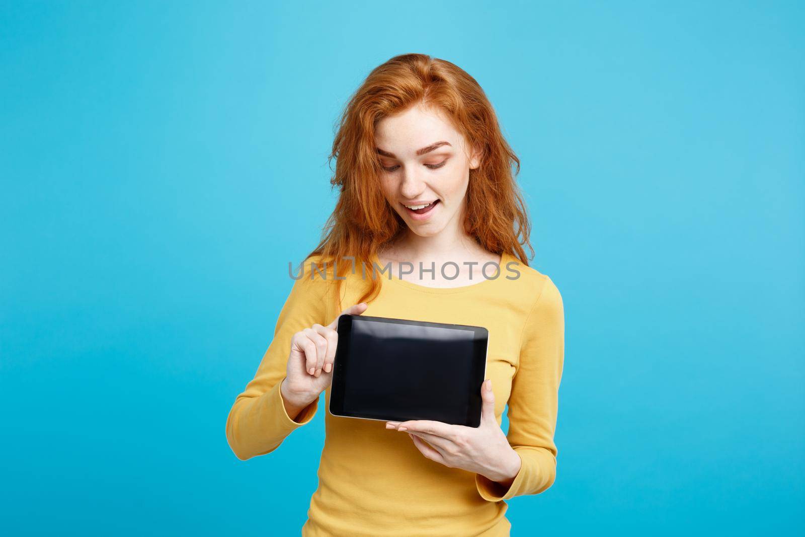 Business Concept - Close up Portrait young beautiful attractive redhair girl smiling showing digital tablet screen on black. Blue Pastel Background.