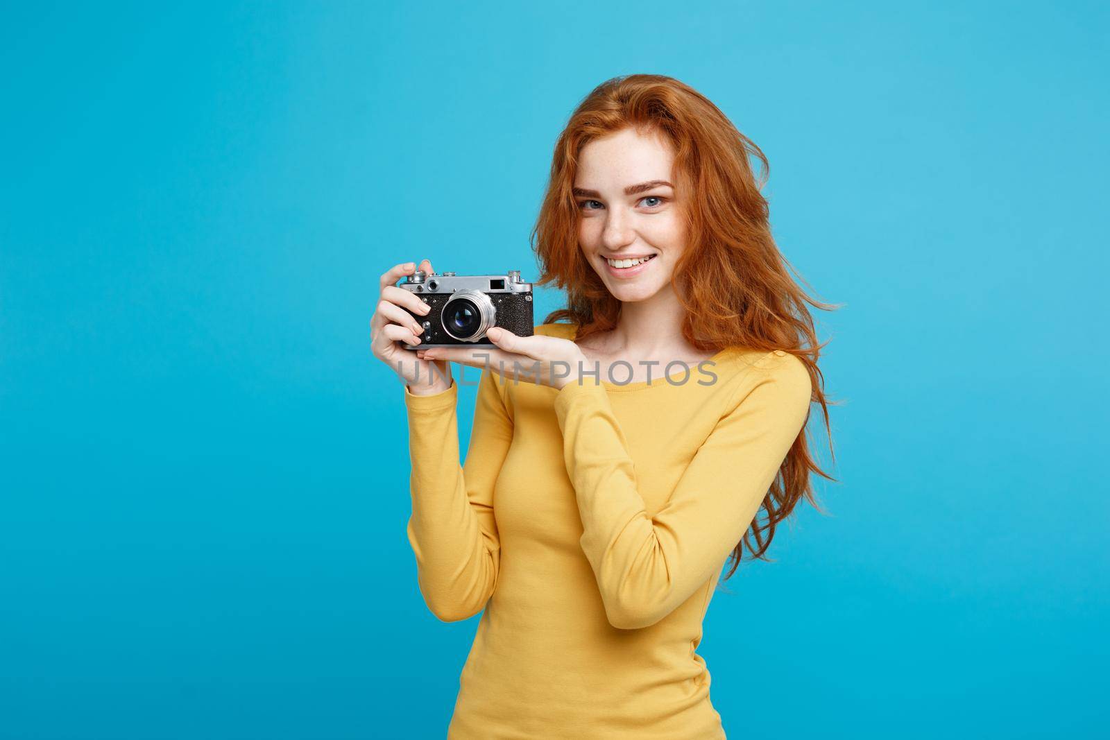 Travel and People Concept - Headshot Portrait of happy ginger red hair girl with playing with vintage camera in happy expression. Pastel blue background. Copy Space.