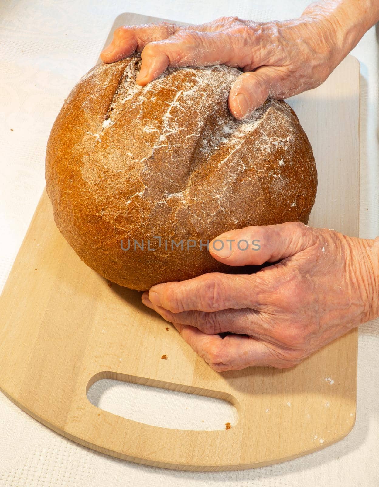 Black fresh round rye bread in the hands of an elderly woman on the kitchen board.   by Puludi