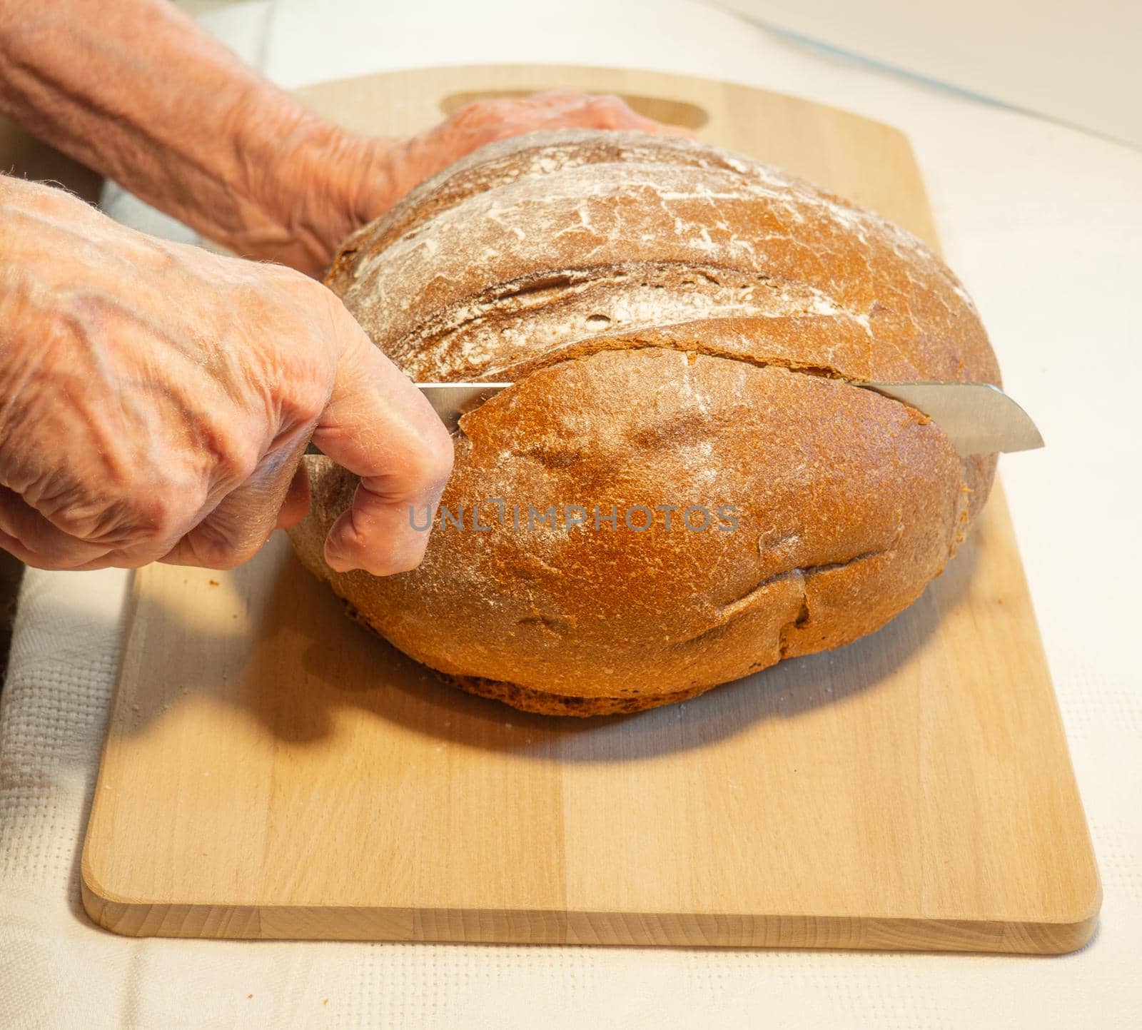 Elderly woman's hands cut round rye bread in flour on kitchen board
