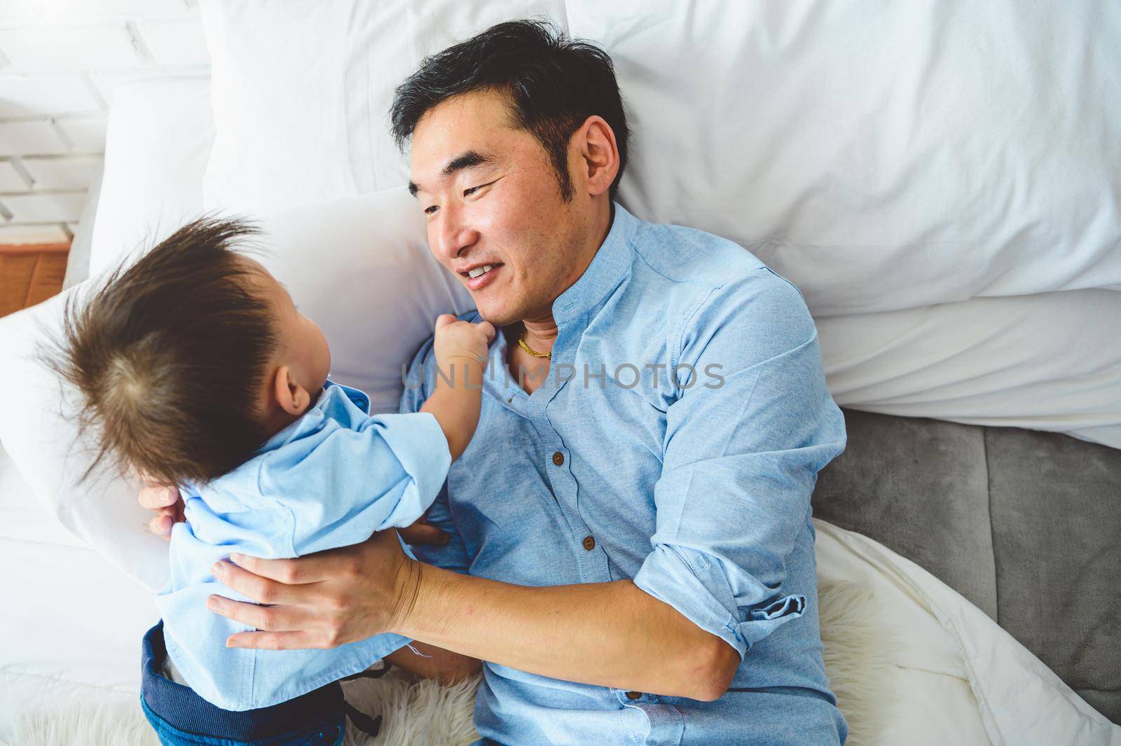 Asian father and son playing together on white bed in bedroom in the morning. Two people having leisure time at home. People lifestyle concept.