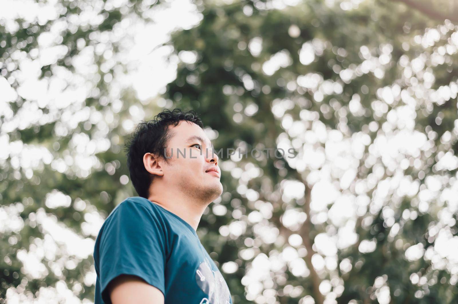 Asian man standing smiling happily under big tree in the public park. Nature and people lifestyle. Healthy single man having success life concept. Copy space