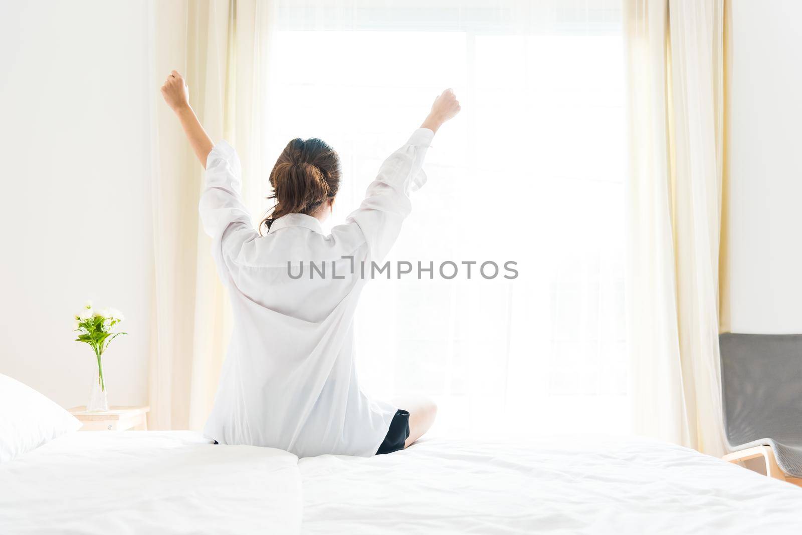 Back view of woman stretching in morning after waking up on bed near window. Holiday and Relax concept. Lazy day and Working day concept. Office woman and worker in daily life theme