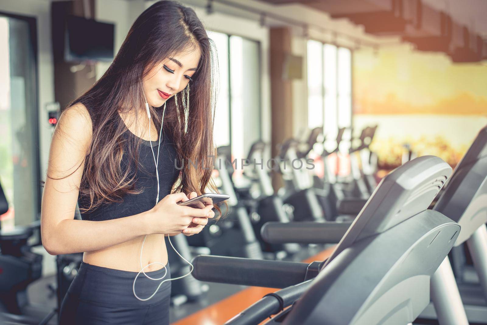 Asian woman using smart phone when workout or strength training at fitness gym on treadmill. Relax and Technology concept. Sports Exercise and Health care theme. Happy and Comfortable mood. by MiniStocker