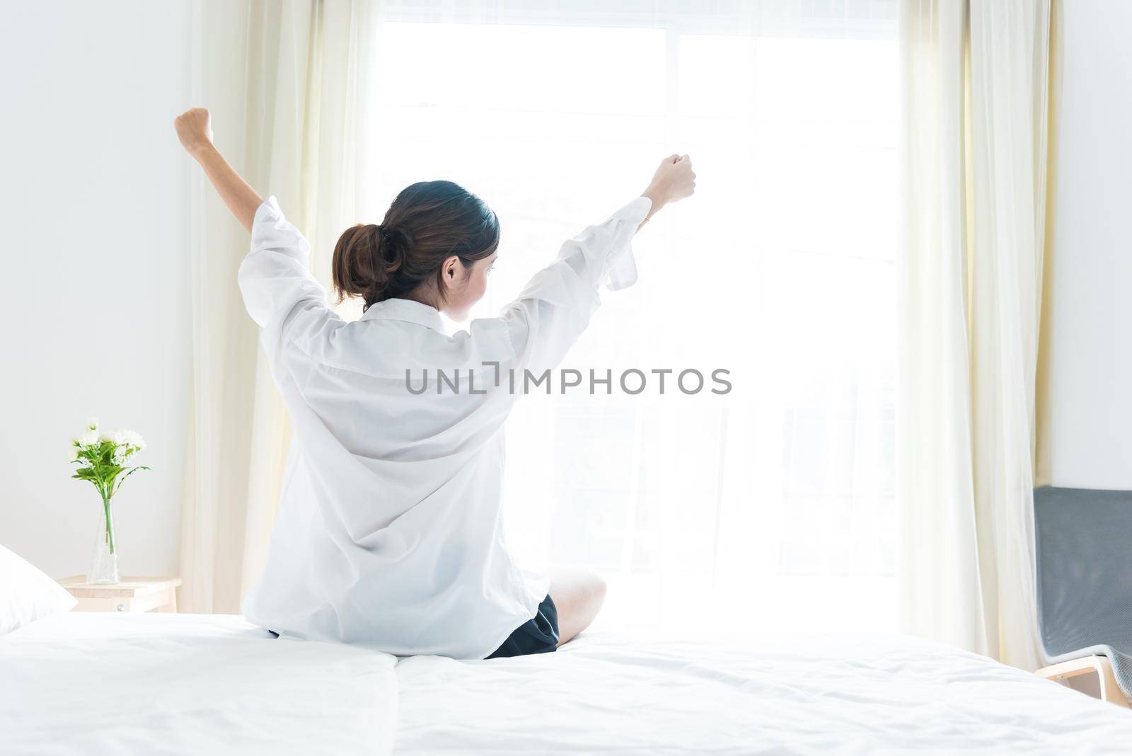 Back view of woman stretching in morning after waking up on bed near window. Holiday and Relax concept. Lazy day and Working day concept. Office woman and worker in daily life theme by MiniStocker