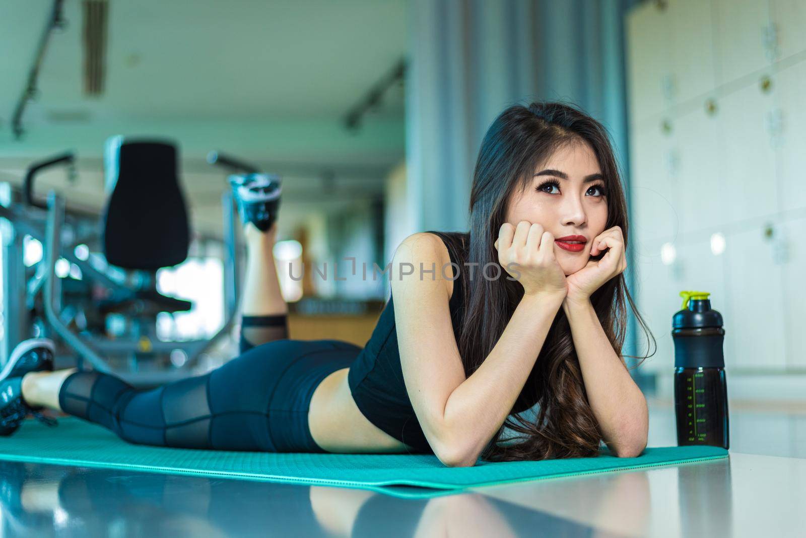 Sports woman lying and relaxing on yoga mat in fitness gym with sports equipment background. Beauty and Workout training exercise concept. Body build up and Strength theme. by MiniStocker