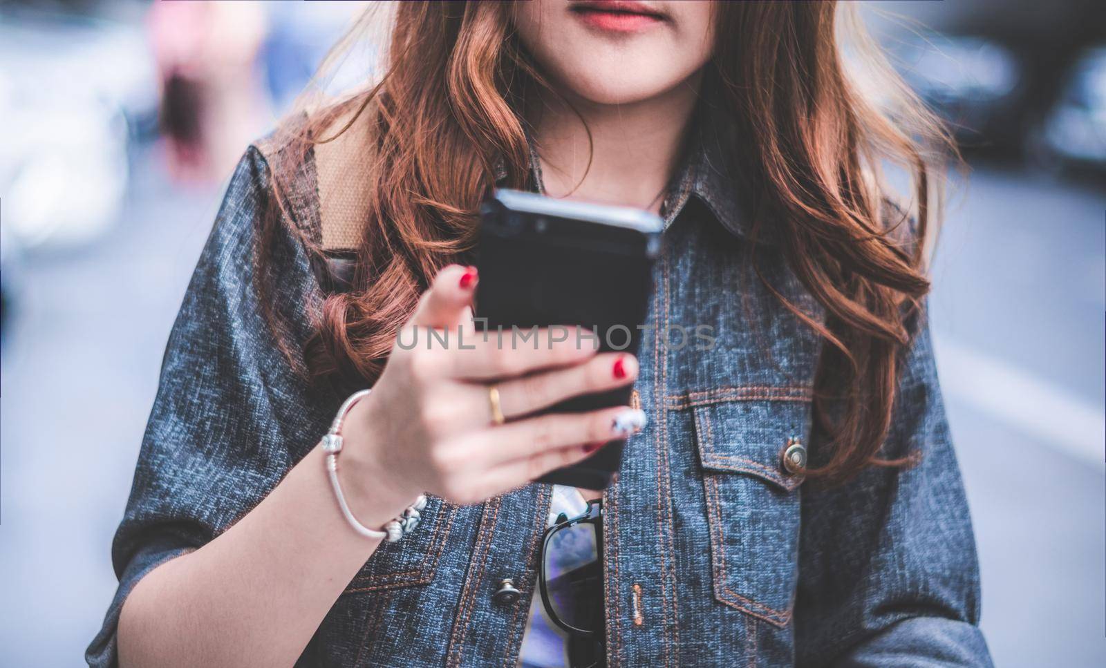 Close up of Beauty woman in jeans jacket using smart phone while walking on street. People and Lifestyles concept. Technology and communication theme. Selective focus on shirt. Blurry background.