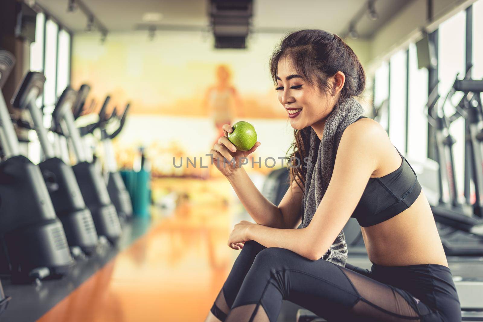 Asian woman holding and looking green apple to eat with sports equipment and treadmill in background. Clean food and Healthy concept. Fitness workout and running theme.