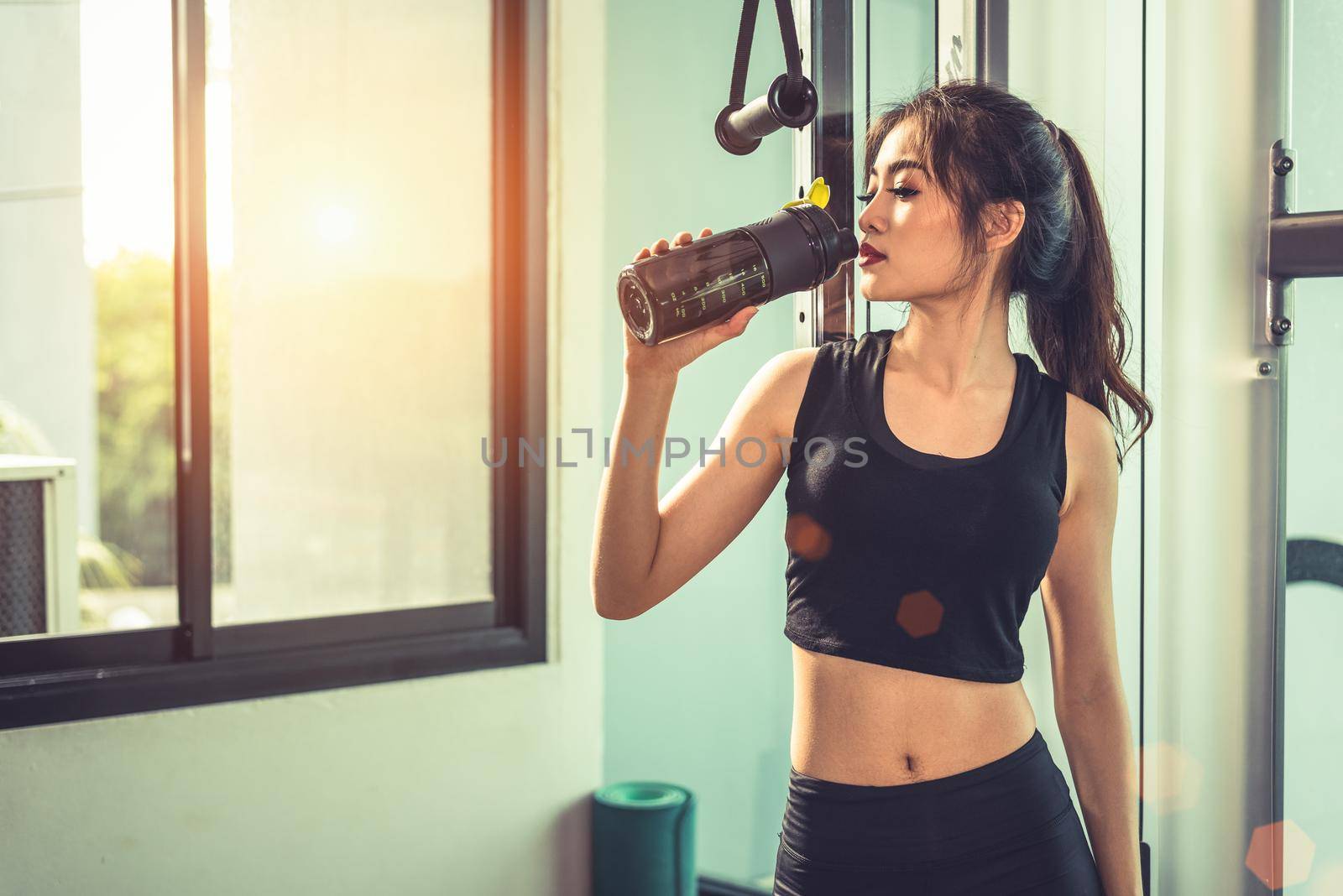 Asian young woman drinking protein shake or water after exercises at fitness gym. Strength training and muscular. Beauty and Healthy concept. Relax concept. Sport equipment and Club center theme. 