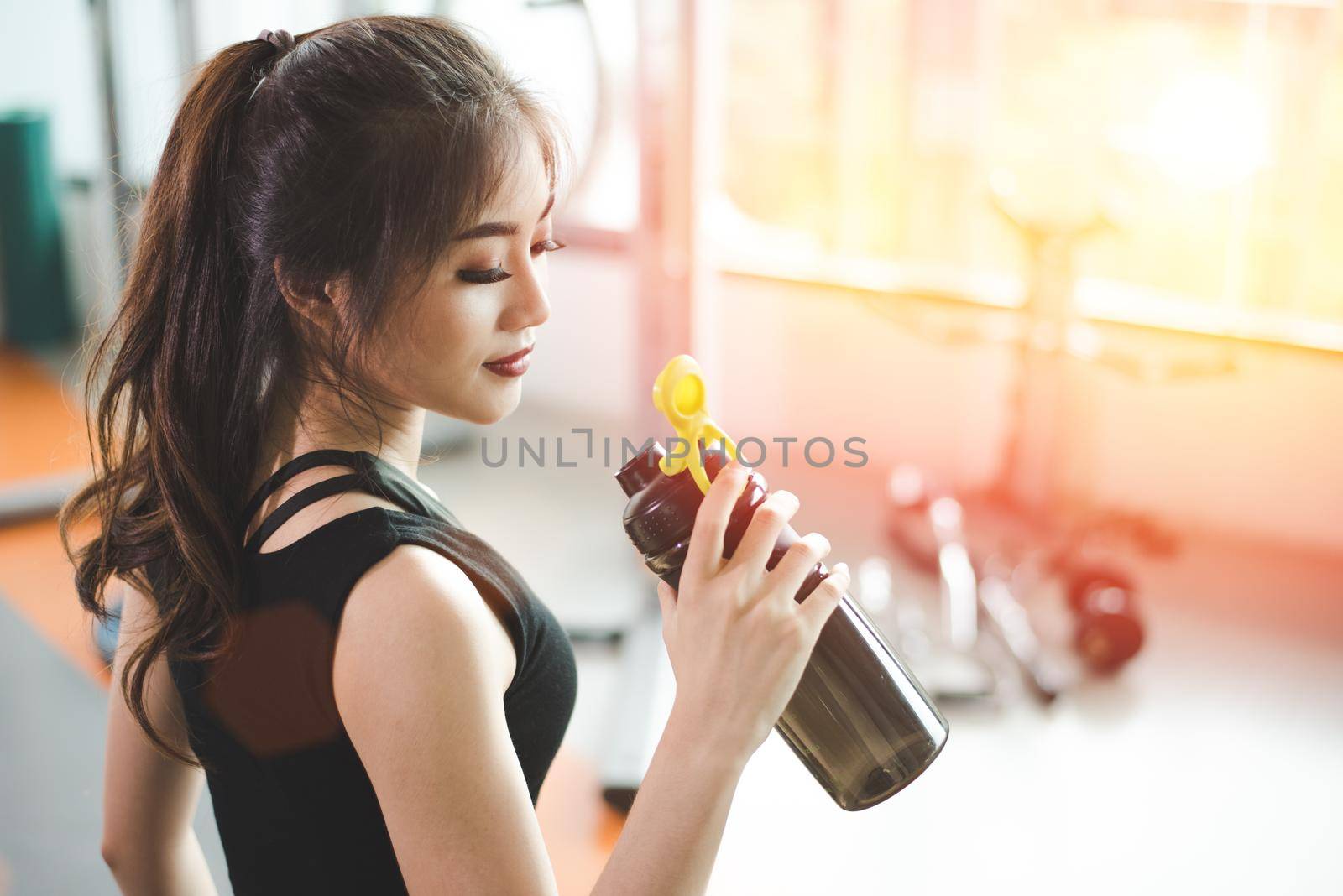Asian beautiful woman drinking protein shake or drinking water in sport fitness training gym. Sports and people concept. Fitness and workout theme.  Sun flare effect