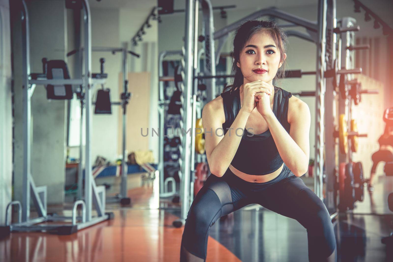 Young Asian woman doing squat workout for fat burning and diet in fitness sports gym with sports equipment in background. Beauty and body build up concept. Sports club and Aerobic theme.