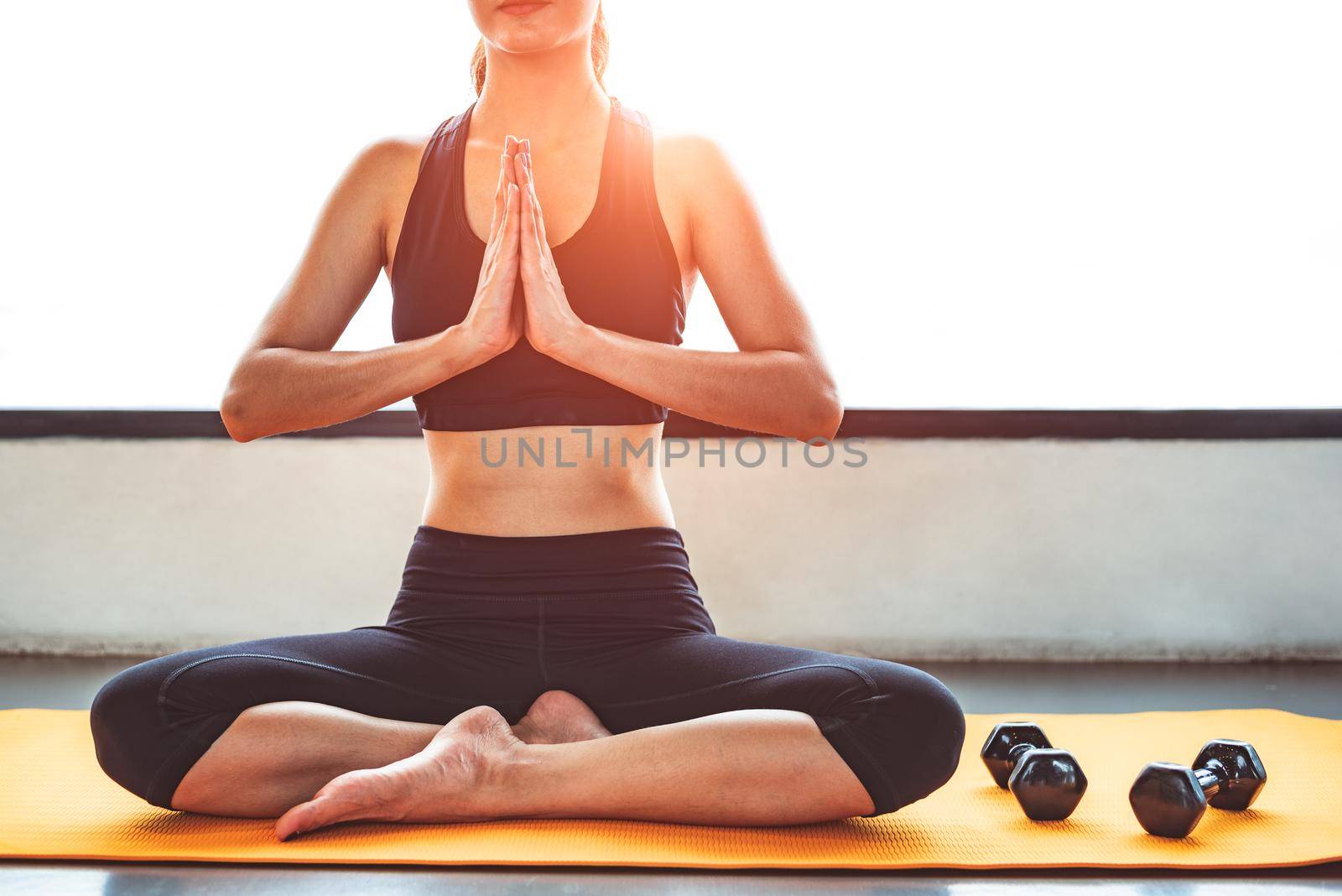 Front view beauty woman doing yoga and raise hand or pay obeisance in fitness workouts training gym center.  Lifestyle sport woman sitting on mat with sport equipment and exercise dumbbells background