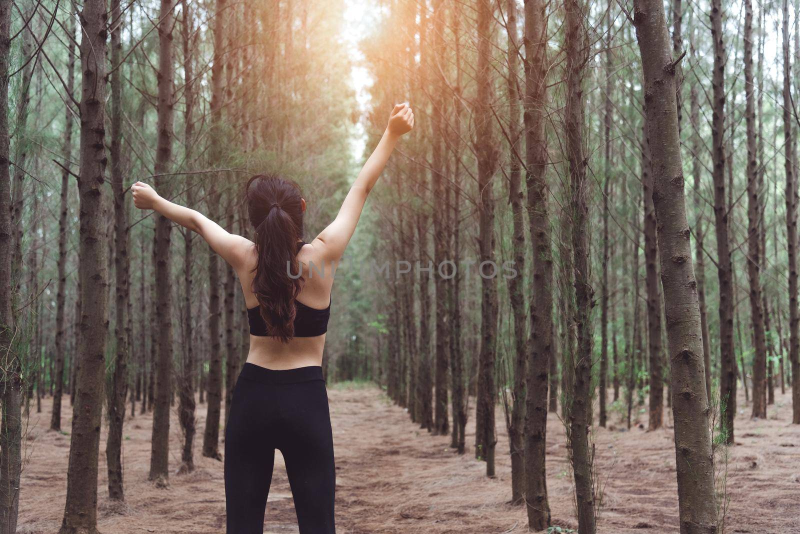 Woman stretching arms and breathing fresh air in middle of pinewood forest while exercising. Workouts and Lifestyles concept. Happy life and Healthcare theme. Nature and Outdoors theme. Back view by MiniStocker