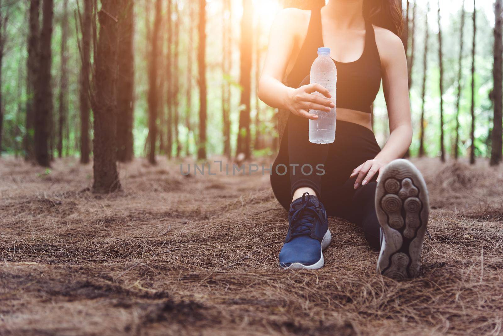 Close up of beauty sport woman resting and holding drinking water bottle and relaxing in middle of forest after tired from jogging. Girl sitting and looking attraction view. Workout lifestyle concept 